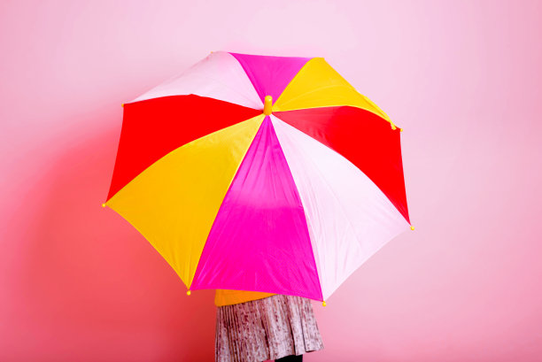 女孩躲在粉红色背景的彩色伞下。图片下载