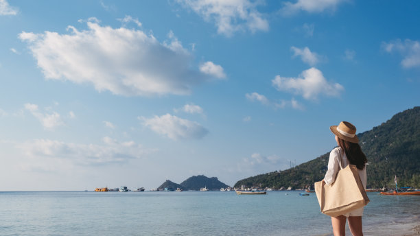 一个带着帽子和包的女人在以蓝天为背景的海滩上散步图片下载