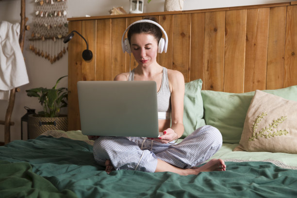早上穿着睡衣戴着耳机躺在床上用笔记本电脑工作的女人。家庭办公室。图片下载