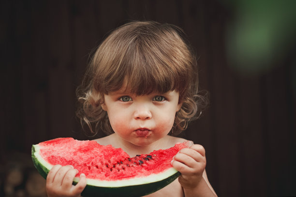 2岁的小男孩抱着一块带种子的成熟红西瓜，咬在天然的木棕色背景上，选择性聚焦，复制空间图片下载