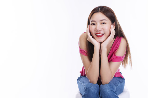 积极的年轻女孩潮人微笑保持她的手下下巴，而坐在孤立的白色背景。友好的女人长头发穿粉红色t恤在工作室享受。图片下载