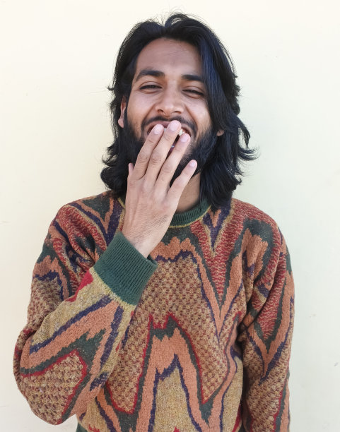 一个英俊的年轻人的肖像笑着用手捂着嘴，工作室拍摄的一个印度年轻人的胡子和长头发-股票照片图片下载