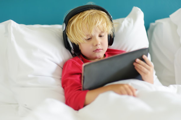 可爱的学前班小男孩早上一起床就戴着耳机在床上用平板电脑看卡通电影。沉迷于电子产品的孩子。图片下载