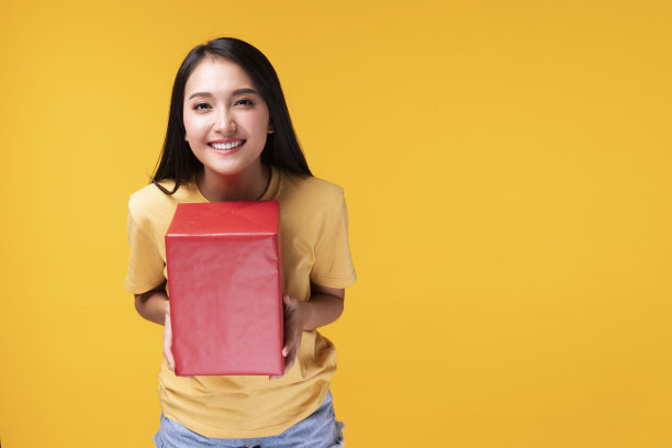 友好的年轻女子手里拿着红色的礼品盒，在黄色的背景下看着相机。兴奋的少年收到惊喜礼物庆祝或周年纪念。图片下载