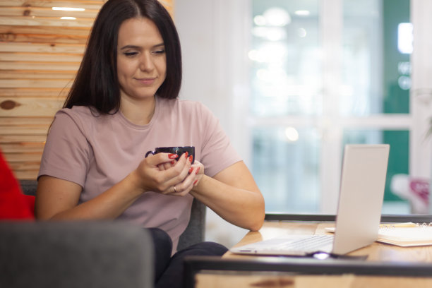 专注的年轻女子在电脑上打字，同时在她的办公室内部喝咖啡休息图片下载