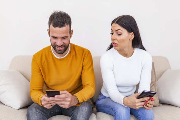 嫉妒的年轻女子用智能手机看着微笑的男友在家里使用智能手机，关系问题概念。不信任的概念图片下载