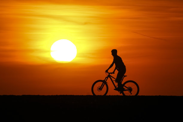 夕阳前的小男孩。大日子在橙色的天空，显示剪影的自行车。天空是橙色的，多云的。绝佳的休闲户外活动。图片下载