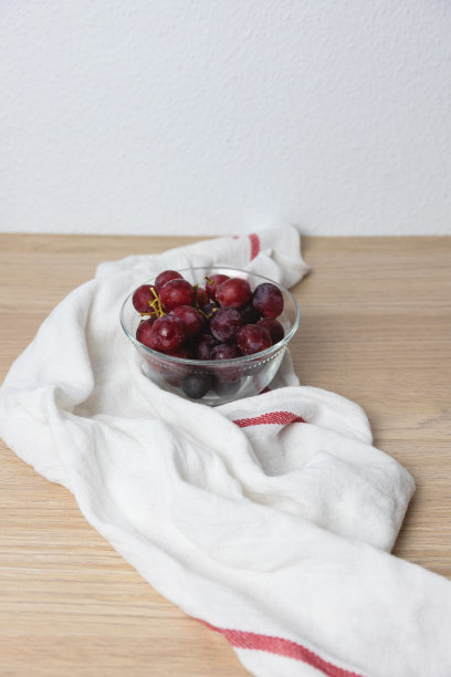 一个盛满红葡萄的小玻璃碗，白色的桌布放在木桌上，白色的背景。甜美的葡萄，红色的果实图片下载