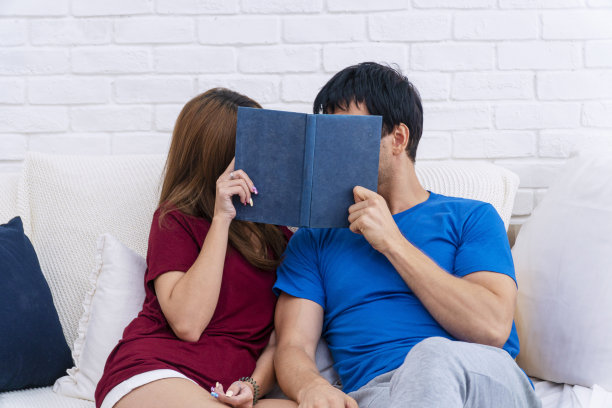 一对浪漫的情侣一起坐在家里的白色沙发上，躲在书后面。英俊的年轻人说话或亲吻他的女朋友封面与书在手。生活方式概念的关系。图片下载