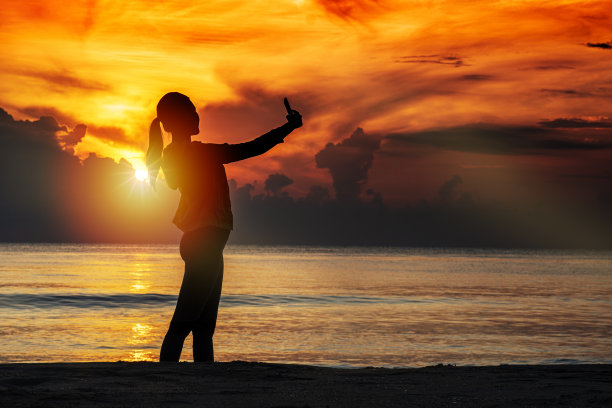清晨日出时，年轻女子在海滩上用智能手机自拍，心情放松。旅游、度假、自然、海洋的概念。图片下载