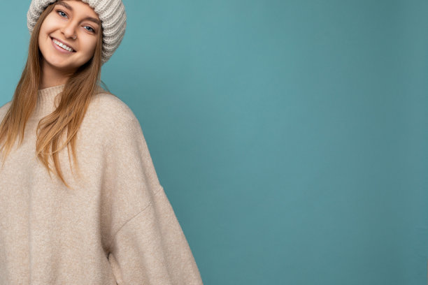 照片迷人的快乐微笑年轻的金发女人孤立在蓝色背景墙穿着米色温暖的毛衣和针织米色帽子看着相机图片下载