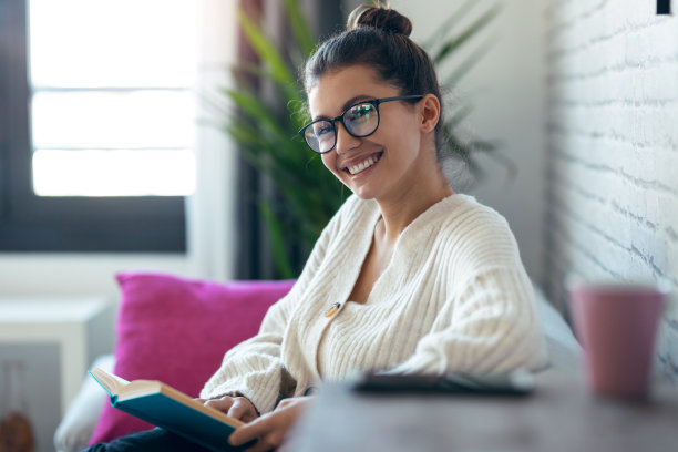 一个漂亮的年轻女人戴着眼镜，坐在家里的沙发上看着相机看书。图片下载