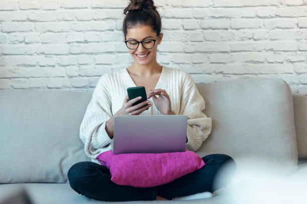 一个年轻漂亮的女人坐在家里的沙发上一边用笔记本电脑工作一边用智能手机发信息。图片下载