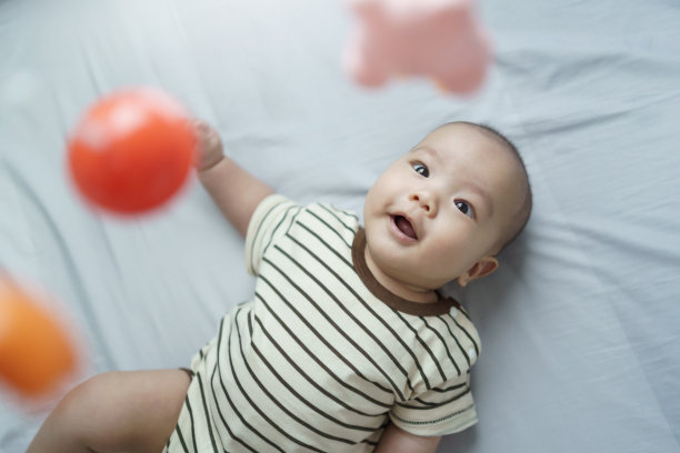 快乐的微笑可爱的亚洲新生男婴躺在床上玩和看移动挂玩具。图片下载