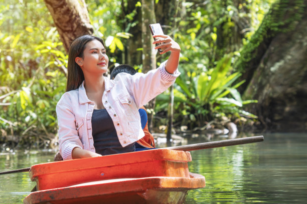 数字技术在假日的概念。享受年轻女子的长发使用智能手机自拍，而坐在船上休闲放松在湖与自然背景。图片下载