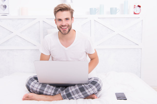 在COVID-19大流行期间，穿着睡衣的乐天派在家里的床上使用笔记本电脑在线工作素材图片