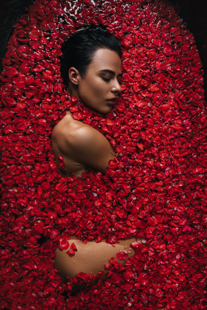 特写，俯视图，美丽的肖像，一个年轻的女人在沐浴与水和浮动的红玫瑰花瓣和玫瑰花蕾的脸。图片下载