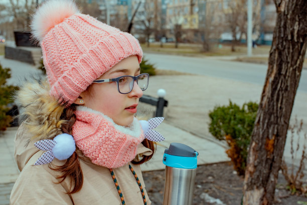 户外肖像可爱的10- 11岁的女孩穿着暖和的夹克。在外面用热水瓶或热水瓶喝茶。戴眼镜的少女。图片下载