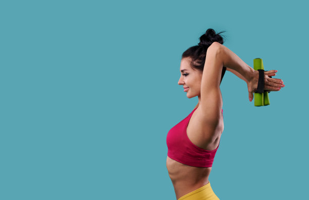 美丽苗条的运动女性在蓝色背景下侧身站立，用哑铃锻炼肱二头肌和手臂的肌肉图片下载