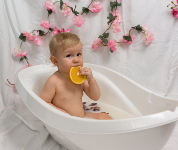 一个小孩坐在浴室里，手里拿着牛奶、橘子和玫瑰花瓣。晚上水疗。一个孩子吃了一片橘子图片下载