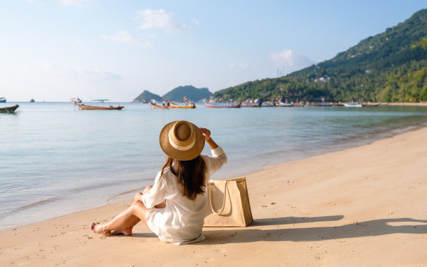 一个带着帽子和包的女人坐在以蓝天为背景的海滩上图片下载