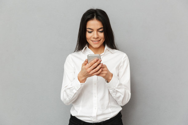 照片内容是一个30多岁的年轻女性穿着正式，手拿智能手机站在灰色的背景下图片下载