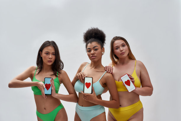 三名多元文化的女性，模特穿着五颜六色的内衣，看着镜头，拿着屏幕上红色心形的智能手机，在灯光背景下摆出孤立的姿势图片下载