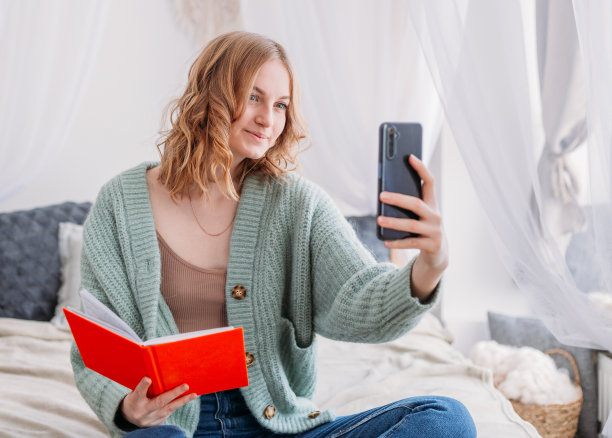 一个漂亮的金发女孩的肖像藏在一个打开的橙色的书和手机后面。快乐的女人在舒适的床上学习或工作。家庭教育或在线交流的概念图片下载