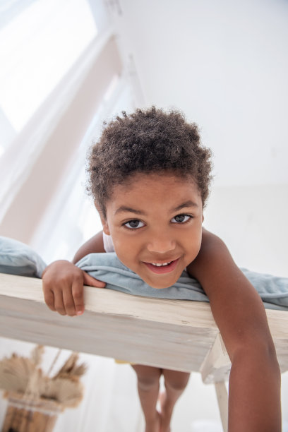 小非洲裔美国男孩玩得很开心，躺在白色的木椅上，挣扎着用他的脚。极简主义的室内设计，蒲苇草，墙壁上的浮木，窗户上的通风门。北欧风格。图片下载