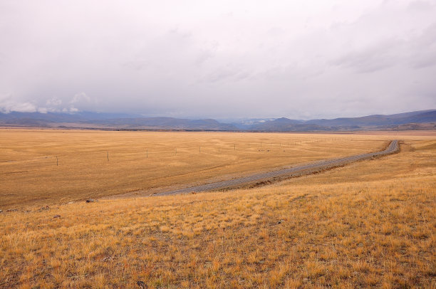 在秋天的大草原上，一条双车道沥青路的转弯，四周环绕着山脉和即将来临的风暴。图片下载