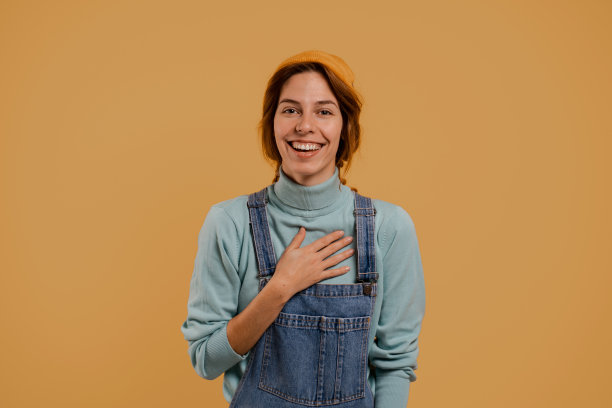 照片中可爱的女农民惊讶地笑着，看起来很开心。穿着牛仔工装裤和帽子，孤立的棕色背景图片下载