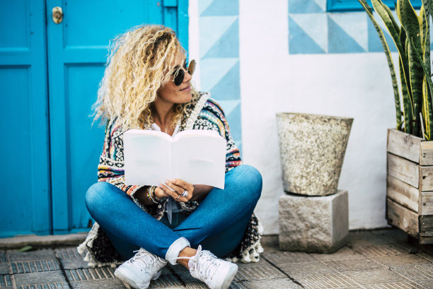 美丽的金发女人读纸书坐在家里外面的地上-蓝色的门和墙壁背景-成年人学习或阅读小说图片下载