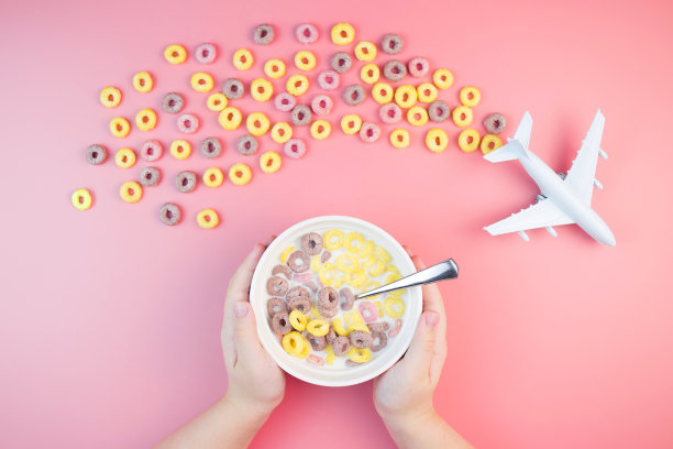 五颜六色的水果玉米圈，碗里有牛奶，孩子们的手，粉红色的背景上有飞机。儿童早餐选择玉米片。图片下载