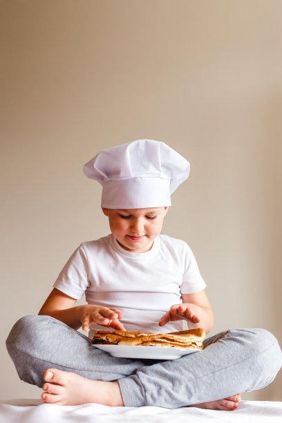 7岁的小男孩穿着白色t恤，戴着白色厨师帽，在白色背景上玩薄饼图片下载