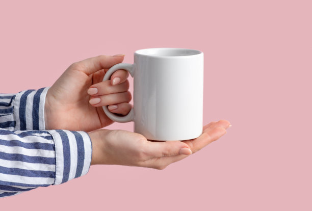 模型的白色咖啡杯或马克杯在女性手中粉红色的背景与复制空间。空白模板为您的设计，品牌，业务。真实的照片。穿着休闲条纹衬衫的女人图片下载