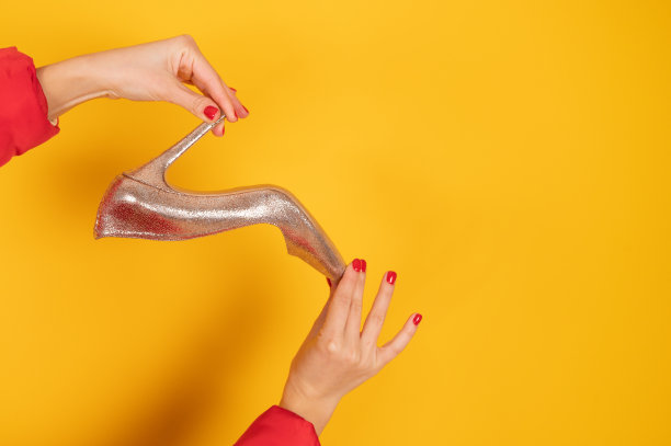剪裁的观点时尚的金色鞋在女性的手与完美的红色美甲。黄色工作室背景与复制空间。伟大的鞋子销售理念图片下载