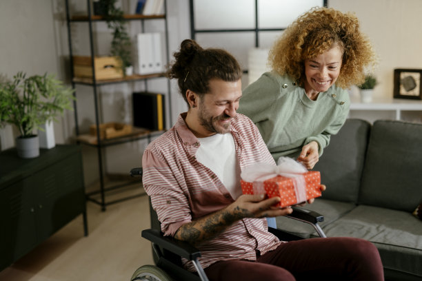 年轻女子在家里用礼物给坐在轮椅上的男友一个惊喜。库存图片图片下载
