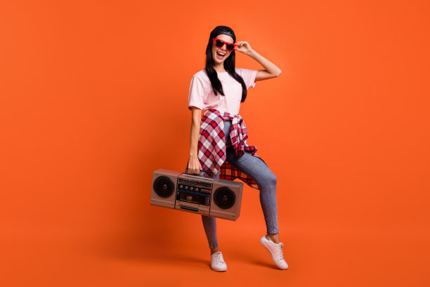 印象深刻的全尺寸照片棕发女士站hold boom box戴帽子眼镜t恤牛仔裤孤立在橙色的背景图片下载