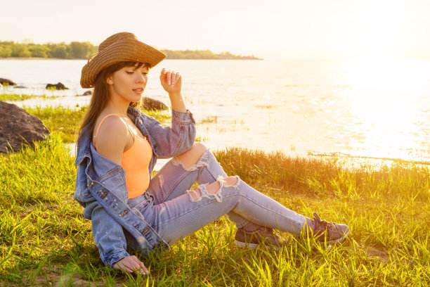 日落时分，穿着牛仔套装，戴着牛仔帽的年轻美女坐在海湾上图片下载