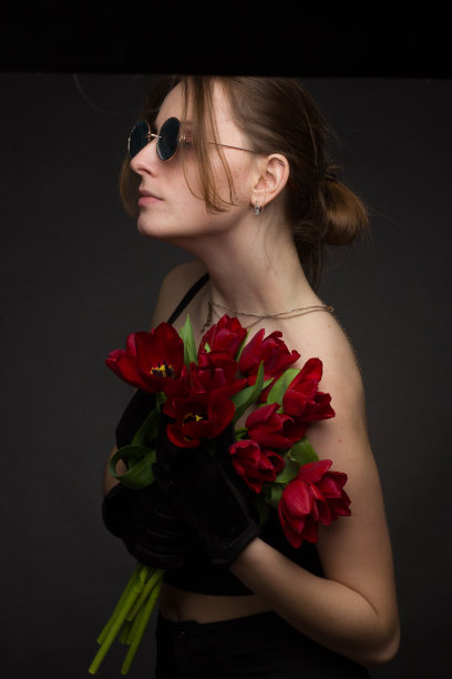 一个戴着墨镜的年轻女子的画室肖像，以黑色为背景，手里拿着一束红色郁金香图片下载