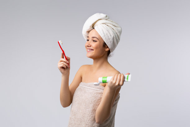 白种女孩与一条毛巾在她的头和包裹在一条毛巾微笑和摆姿势与牙刷和粘贴在一个灰色的背景与侧空间。早上程序。图片下载