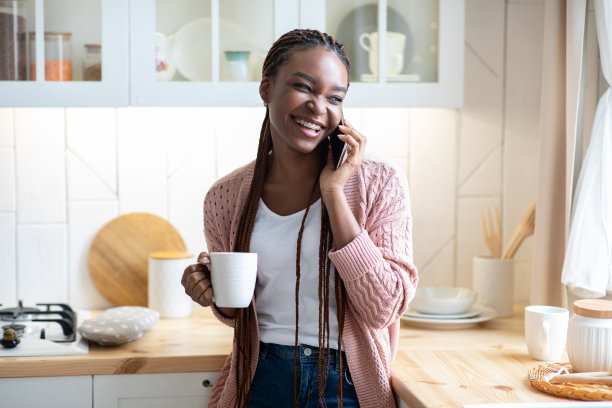 快活的黑人妇女在厨房里一边打电话一边喝咖啡图片下载