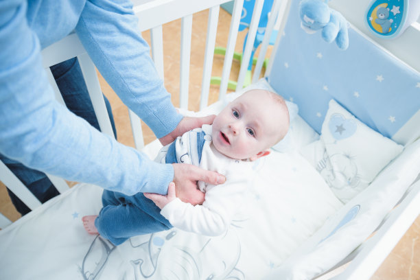 父亲穿着蓝色的衣服，他的孩子躺在他的白色婴儿床里图片下载