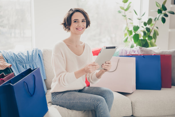正波波头女孩的侧面照片坐在沙发上拿着平板电脑网上购物袋在室内远程购买图片下载