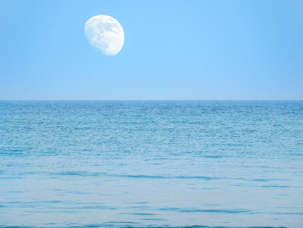 月亮在蓝天上悬在海面或海洋的上空。图片下载