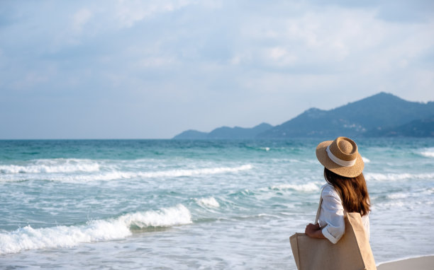 一个带着帽子和包的女人在以蓝天为背景的海滩上散步图片下载