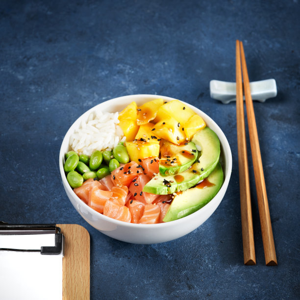 快速健康午餐，三文鱼戳碗生鱼沙拉亚洲流行食品，芒果，大豆毛豆，米饭，鳄梨，盛在碗里，筷子，蓝色的深色背景。副本的空间。有选择性的重点。图片下载