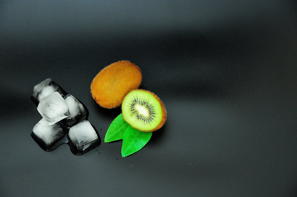 两个成熟的猕猴桃，整个和一半，旁边的冰块和叶子在黑色的背景。图片下载