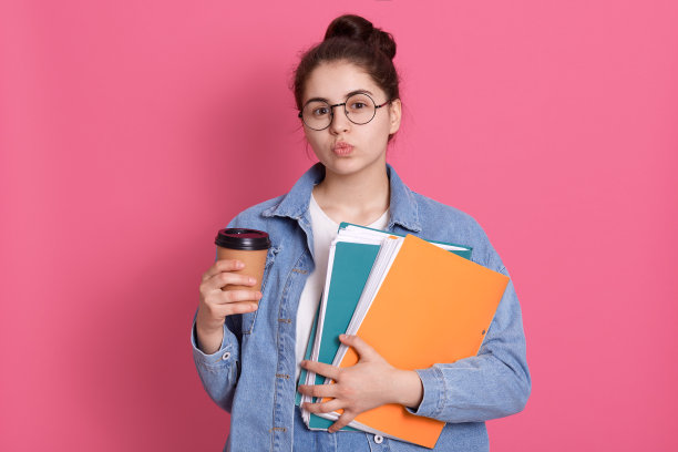 可爱的黑头发女学生，嘴唇圆润，在学校和别人调情，戴着圆圆的眼镜，拿着彩色的纸夹，一次性咖啡或卡布奇诺。图片下载