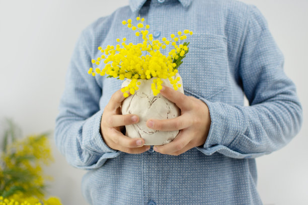 为母亲节、3月8日或生日制作陶瓷工艺品。一个孩子拿着一个手工花瓶，上面插着黄色的含羞草花。简单的礼物的想法。图片下载